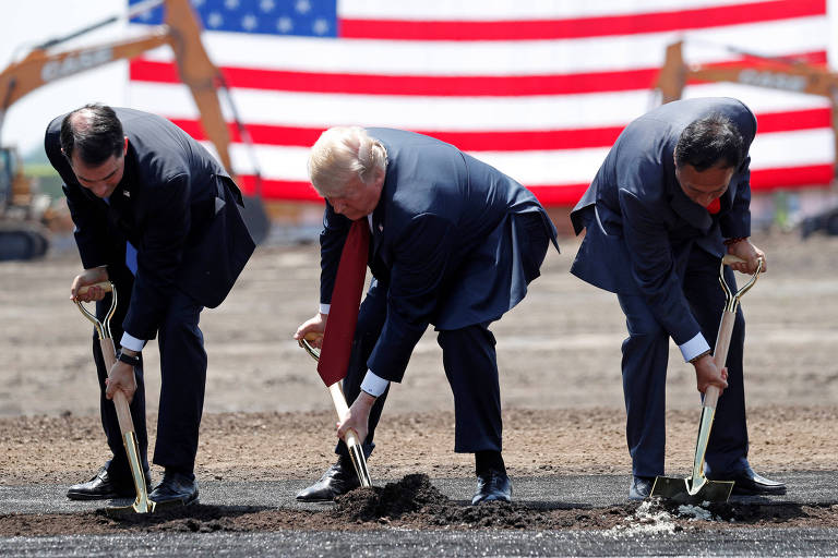 Em frente a uma grande banderia dos EUA, com listras vermelhas e brancas e um quadrado azul com estrelas brancas, três homens de terno se agacham em posições meio ridículas para cavar o solo com pás