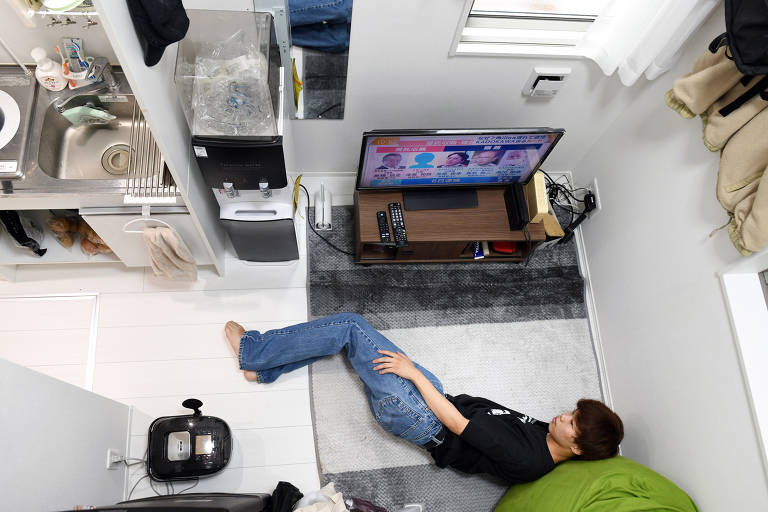 Jovem japonês de camiseta preta e calça jeans assiste televisão deitado no chão em seu pequeno apartamento em Tóquio, no Japão