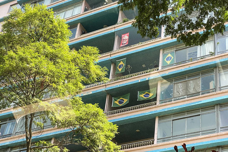 Imagem mostra bandeiras do Brasil e do Lula penduradas em sacadas de apartamentos de um prédio residencial. Há árvores na frente do prédio.