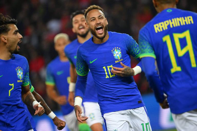 Fotografia colorida de Neymar (centro) celebrando gol do Brasil. Ele veste a camisa 10 de cor azul da seleção e aparece com a boca aberta e sorrindo. Ao lado direito, de costas para a câmera, está o Raphinha, com a camisa 19
