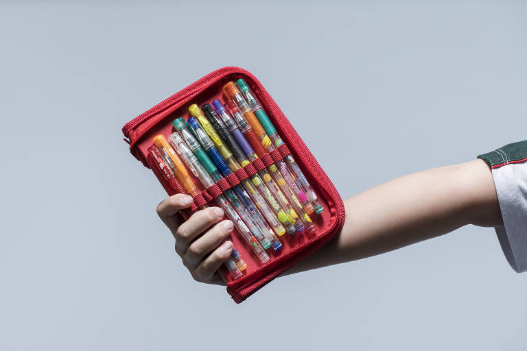 Criança segura com uma das mãos um estojo vermelho com canetas coloridas
