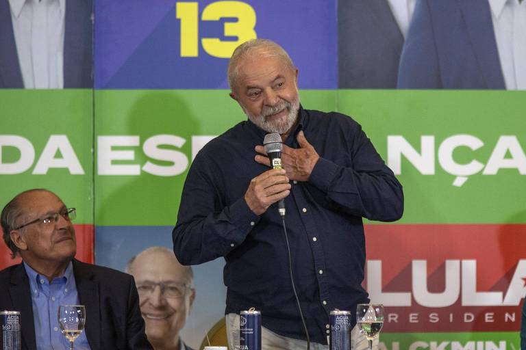 Ex-presidente Luiz Inácio Lula da Silva, de pé com o microfone na mão e camisa azul escura, com o vice na chapa, Geraldo Alckmin, sentado ao seu lado