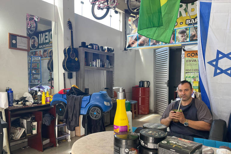Homem de meia-idade bebe chimarrão sentado atrás de uma mesa. Ao fundo, se enxergam as cadeiras da barbearia e bandeiras de Brasil e Israel.