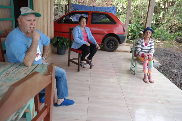 Homem de meia idade de boné está escorado em mesa em frente a uma casa de madeira. Ao fundo, sua esposa e sua mãe. Atrás delas, há um carro popular vermelho estacionado.