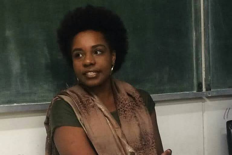 Professora negra esta sentada em frente ao quadro verde. Ela usa uma calçapreta, camiseta verde e um cachecol marrom. Seu cabelo é afro e ela está olhando para o lado. 