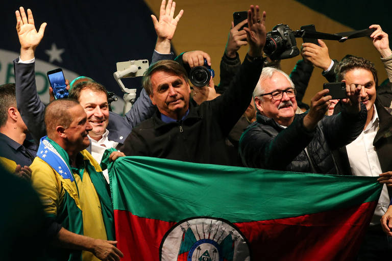 O presidente Jair Bolsonaro (PL) acena atrás de bandeira do Rio Grande do Sul em evento com apoiadores na cidade de Pelotas
