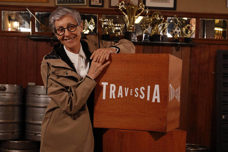 Atriz Cássia Kis aparece de casaco comprido marrom atrás de uma caixa com o logotipo da novela Travessia