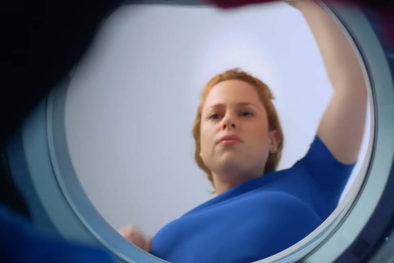 Reprodução colorida de vídeo de campanha de Omo. Uma mulher jovem, branca e ruiva, com camiseta azul, está de frente para a câmera abrindo uma máquina de lavar, com a câmera dentro do eletrodoméstico