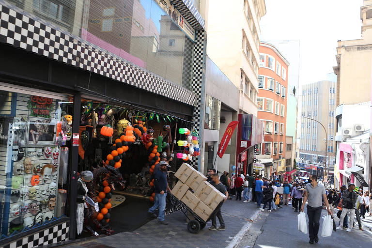 Imagem mostra prédios e comércio na 25 de Março. Na rua, pessoas caminham com sacolas na mão.