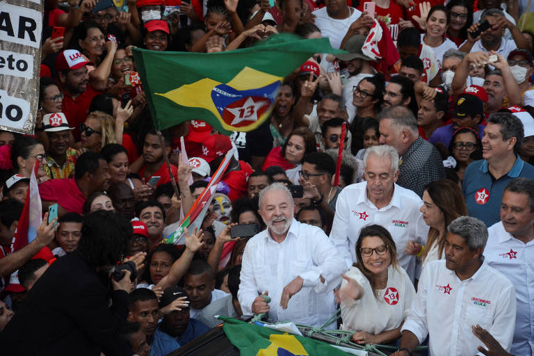 Lula está de camisa branca em meio à multidão de apoiadores usando vermelho e com bandeira do Brasil ao fundo