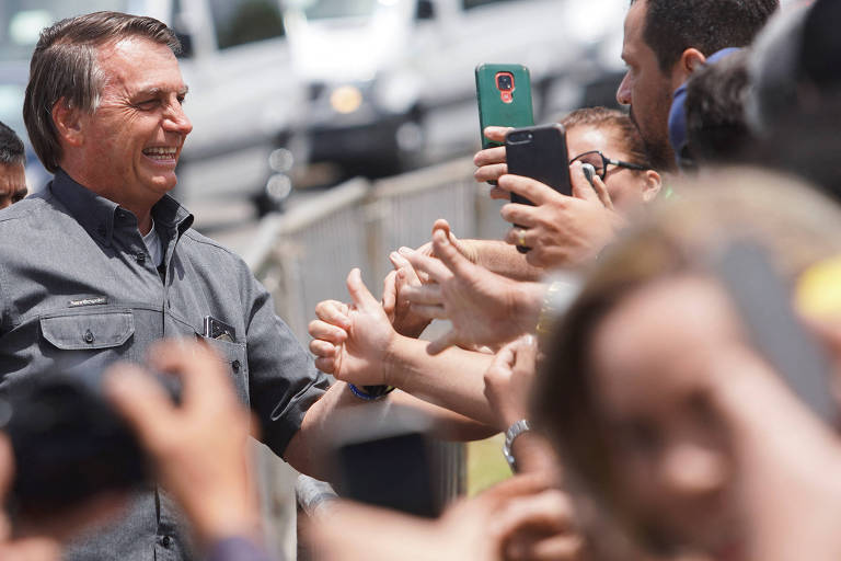 Bolsonaro aparece no canto esquerdo da foto, de camisa cinza escuro, sem paletó, sorrindo, enquanto mãos de várias pessoas tentam cumprimentá-lo e algumas seguram celulares para fazer fotos