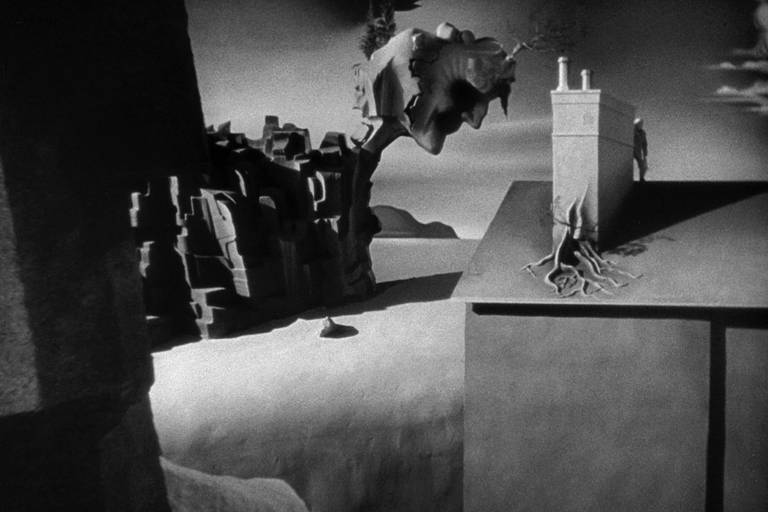 cena em preto e branco mostra uma paisagem cenográfica criada pelo artista surrealista Salvador Dalí, com a figura de um rosto de homem ao fundo derretendo, uma casa com uma chaminé e raízes saindo de sua base e uma planície vazia