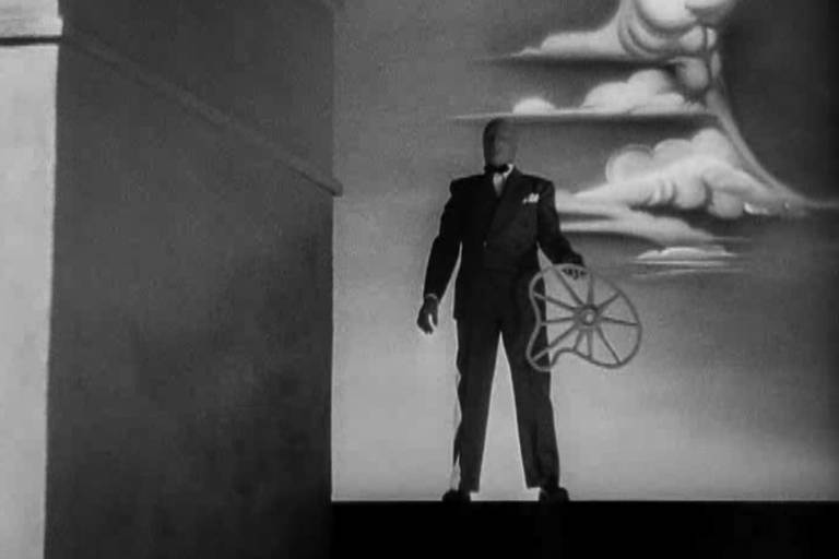 Cena mostra homem sobre telhado de casa ao lado de uma chaminé com o rosto coberto por um tecido e vestido com terno, segurando uma roda de carroça torta com nuvens ao fundo
