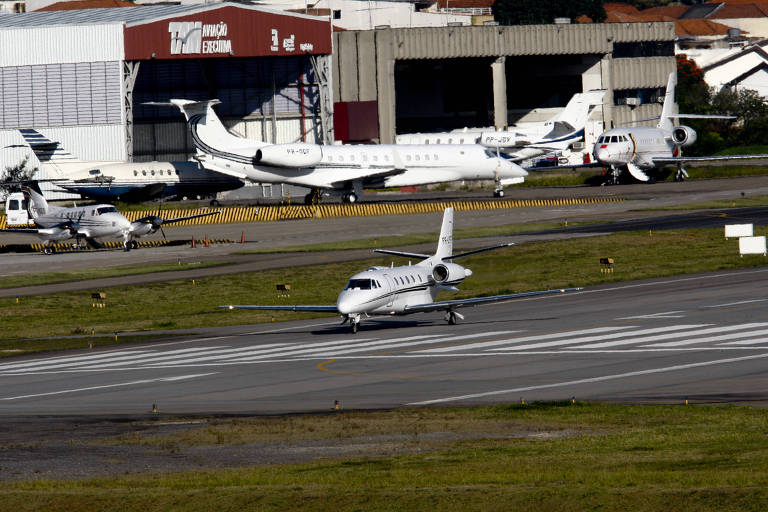 Imagem mostra jatinho em pista de aeroporto. Ao fundo, alguns outros aviões estão parados em hangares.