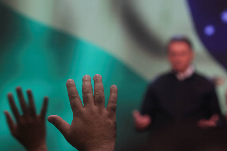 Mãos de fieis erguidas em primeiro plano; atrás, Bolsonaro diante da bandeira do Brasil em culto com Silas Malafaia