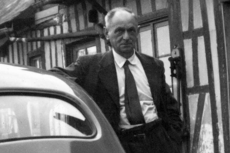 Foto preto e branco mostra homem de terno e gravata, encostado num carro em frente a uma casa típica normanda, com vigas de madeira cruzadas na fachada