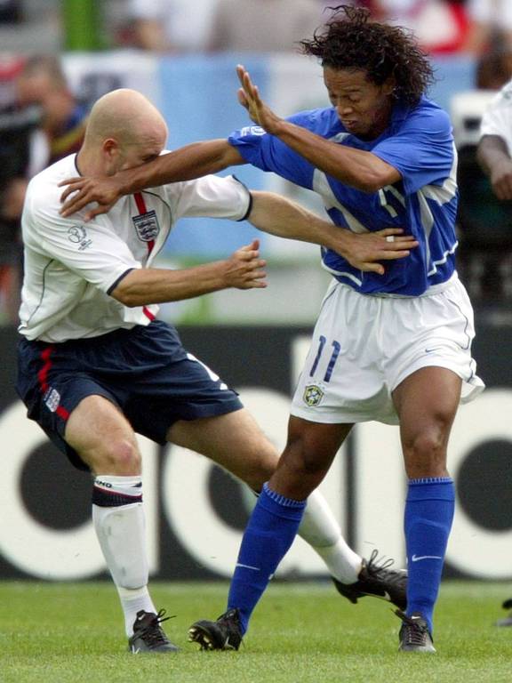 Ronaldinho, de camisa azul, meiões azuis e calção branco, coloca o braço direito no rosto de Danny Mills, de camisa branca, calção azul escuro e meiões brancos, em lance na partida em que o Brasil ganhou por 2 a 1 da Inglaterra na Copa do Mundo de 2002, em Shizuoka, no Japão
 