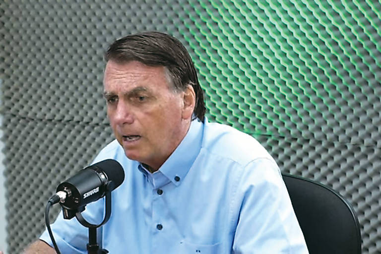 Imagem mostra o presidente Jair Bolsonaro durante participação de um podcast. Ele é um homem branco que veste uma camisa azul clara e fala no microfone