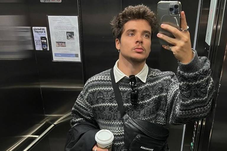 Em foto colorida, homem de camisas listradas de mangas compridas faz selfie no elevador