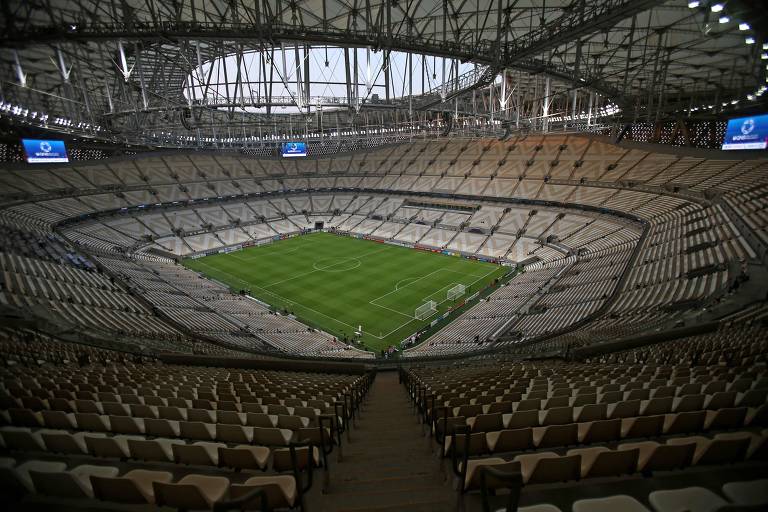 Estádio tem gramado verde no centro e arquibancada vazia