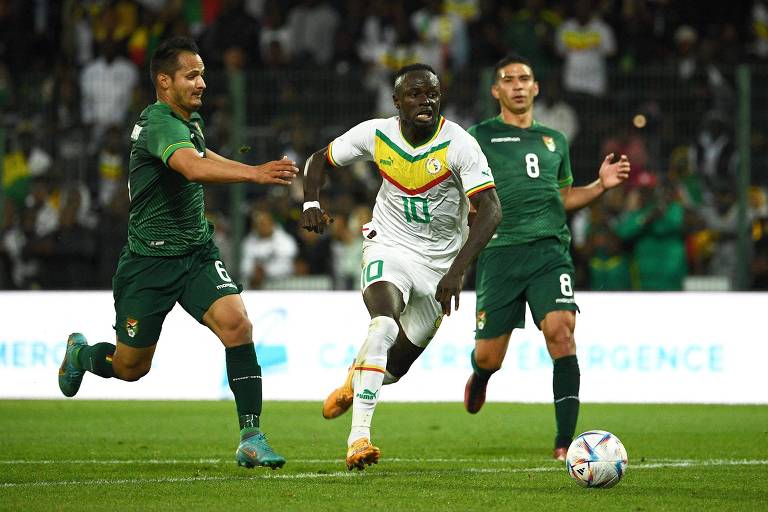 Marcado por Justiniano, que corre ao seu lado de uniforme verde, Mané, com o número 10 estampado no peito da camisa branca com detalhes em amarelo, verde e vermelho, avança com a bola em amistoso do Senegal contra a Bolívia em Orleans, na França