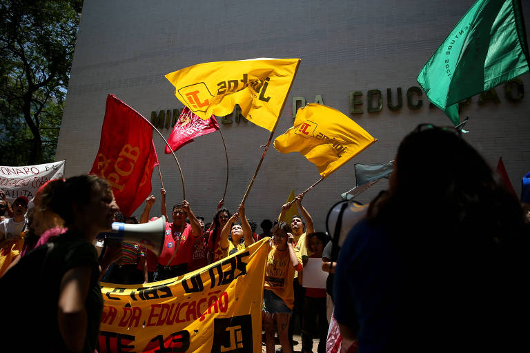 grupo de manifestantes em frente ao prédio do MEC, onde é possível ler parte da palavra educação. Há bandeiras amarelas, vermelha e verde