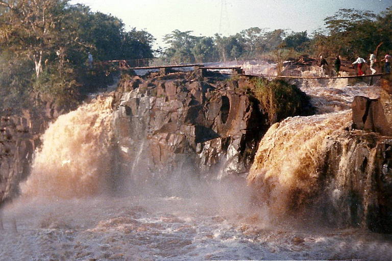 Cachoeira desce por pedras enquanto algumas pessoas andam por uma ponte em cima da queda d'água