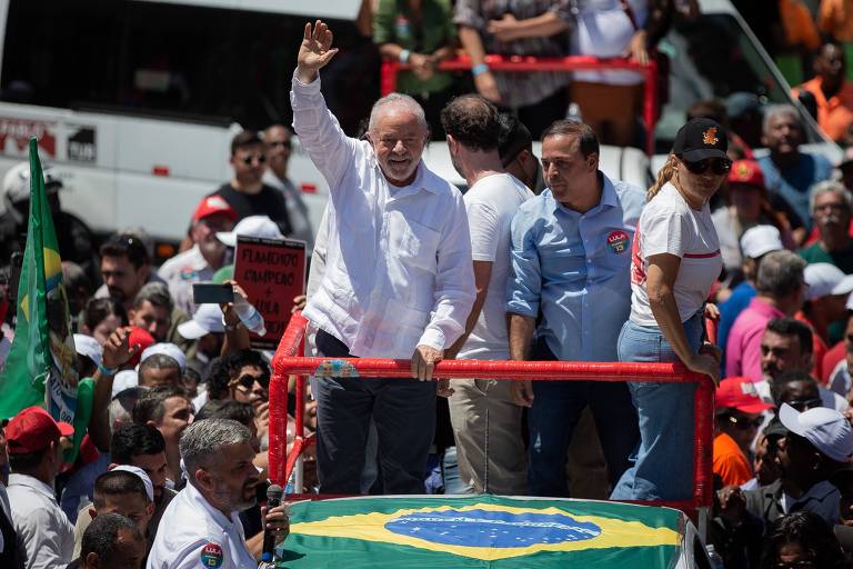Lula é um homem branco, idoso, calvo, com cabelo e barba grisalhos, nariz um pouco achatado e orelhas grandes. Está ao centro da imagem, em um carro com a bandeira do Brasil, e Lula levanta os braços em tom de vitória. Ao redor do carro, muitos apoiadores, militantes e fotógrafos