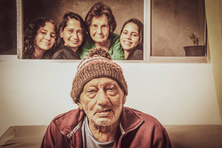 Imagem colorida mostra Olavo Nogueira, um homem de 86 anos. Ele aparece do tronco para cima, veste uma blusa vermelha e um gorro marrom. Ao fundo, em uma janela, estão quatro mulheres, todas elas sorrindo.