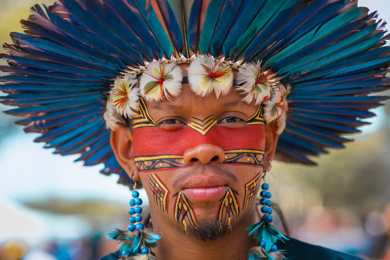 Fotografia colorida em close do indígena Tukumã Pataxó, com desenhos vermelhos e amarelos pelo rosto e cocar azul na cabeça