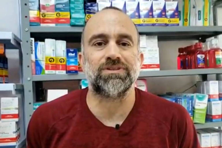 Homem de cabelo curto, barba grisalha, de camiseta vermelha na frente de prateleira de medicmentos
