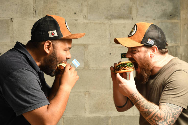 Dois homens estão frente a frente, segurando e mordendo hambúrgueres; ambos têm barbas, usam bonés e camisetas