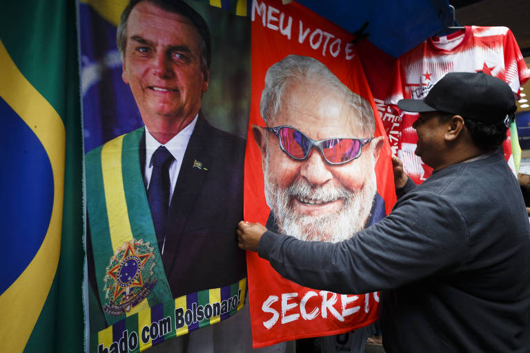 Imagem mostra homem segurando toalha vermelha com rosto de Lula. Ao lado, há uma toalha com a foto de Jair Bolsonaro.