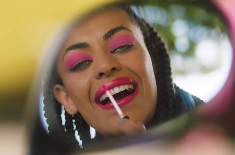 Fotografia colorida de mulher passando gloss na boca, vista pelo espelho retrovisor de um carro. A mulher é negra, tem cabelos pretos, longos e trançados e usa uma maquiagem rosa nos olhos.