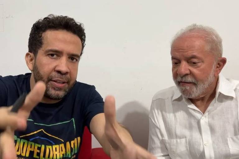 O deputado está de camisa preta e gesticula para a câmera, com uma caneta na mão; Lula observa, vestindo camisa branca