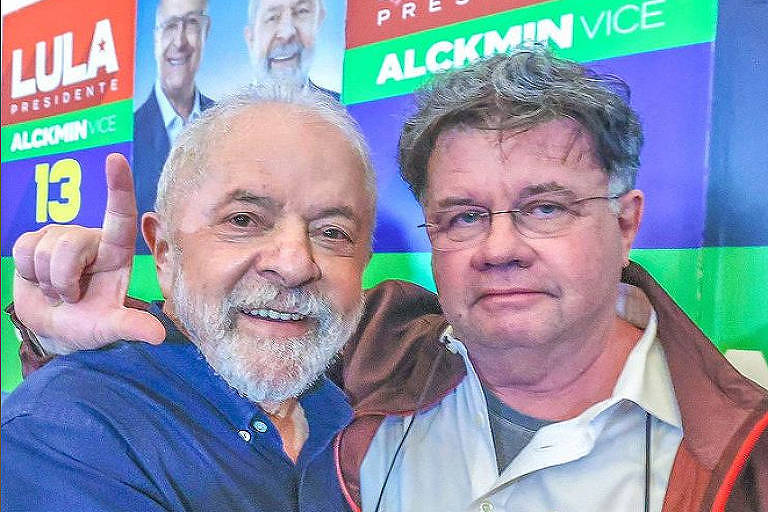Lula, homem branco de cabelo e barba grisalhos, está de camisa azul e abraçado a Marcelo Madureira, homem branco de cabelo grisalho, de camisa branca e casaco aberto. Marcelo faz o L com a mão direita. Ao fundo, painel com imagens de Lula e Geraldo Alckmin, vice de Lula