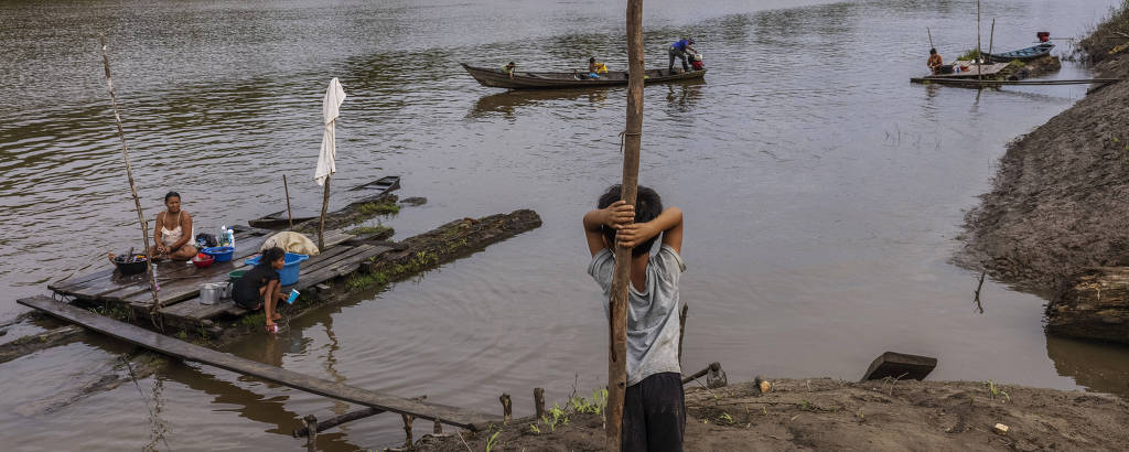 Criança se apoia em poste na beira de um rio, enquanto dois adultos lavam roupas na beira da água