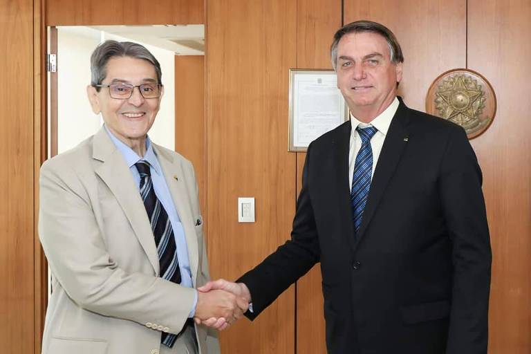Ex-deputado Roberto Jefferson e o presidente Jair Bolsonaro (PL) juntos, em imagem divulgada em setembro de 2021 pelo PTB, partido de Jefferson. Ambos estão de terno e dão a mão, enquanto olham para a camaera