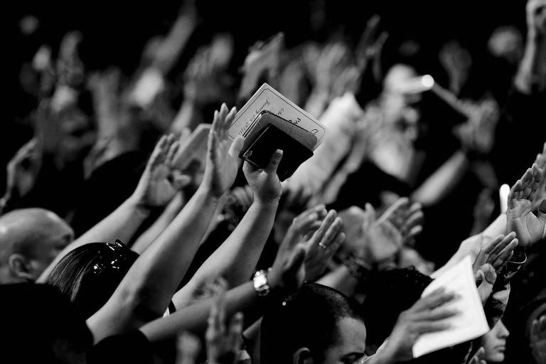 Fiéis evangélicos oram em um culto com os braços erguidos, alguns segurando bíblias