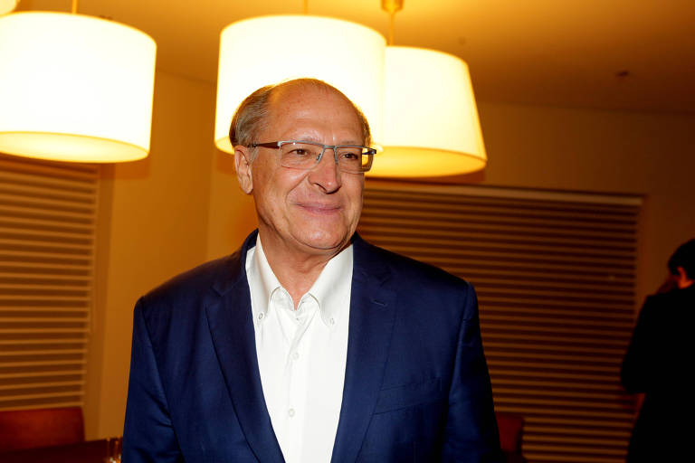 Geraldo Alckmin, homem branco, usa óculos e veste camisa social branca e paletó de tom azul. Ao fundo, lustres; ele está em sala com iluminação amarela