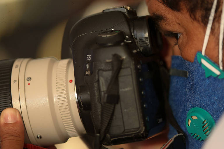 Um homem olha pela lente de uma câmera fotográfica. Ele usa máscara de proteção no rosco e ajusta o foco com a mão