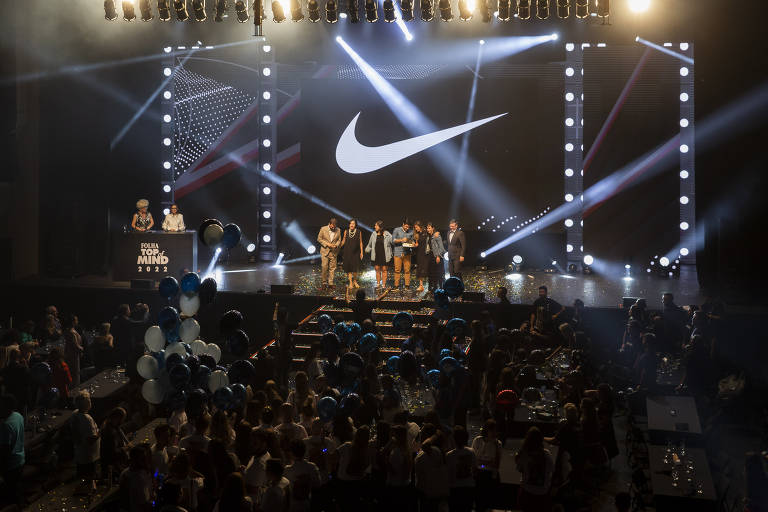 Imagem mostra plateia de frente a palco. Um telão exibe o logotipo da Nike.