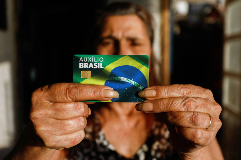 Mulher segura nas mãos cartão bancário estampado com o símbolo do programa federal Auxílio Brasil
