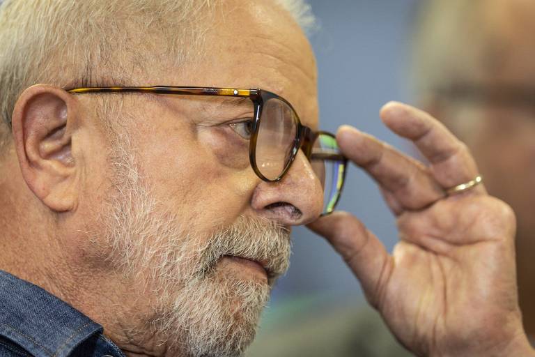 Lula está com semblante sério e olha para a frente. Ele ajusta os óculos em seu rosto com uma das mãos. O ex-presidente é um homem branco e tem barba e cabelos brancos