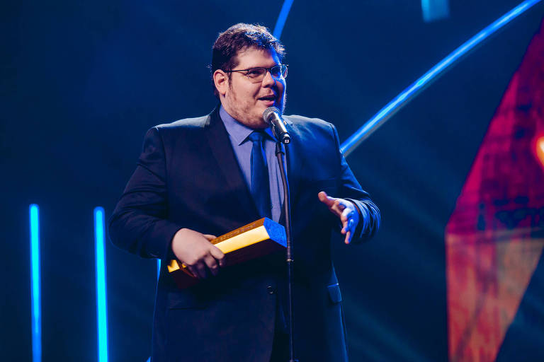   Em foto colorida, homem de terno azul marinho discursa ao microfone durante premiação