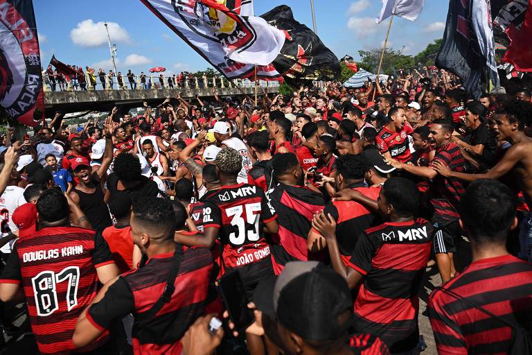 Fotografia colorida mostra multidão de torcedores do time de futebol carioca Flamengo. Eles utilizam camisa listrada nas cores vermelho e preto e carregam bandeiras do time. 