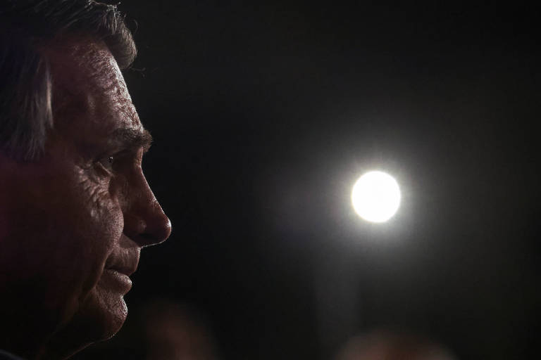 A imagem escura mostra o rosto de Bolsonaro, um homem branco, no canto, de perfil. Diante dele, um ponto de luz aparece em meio ao fundo escuro