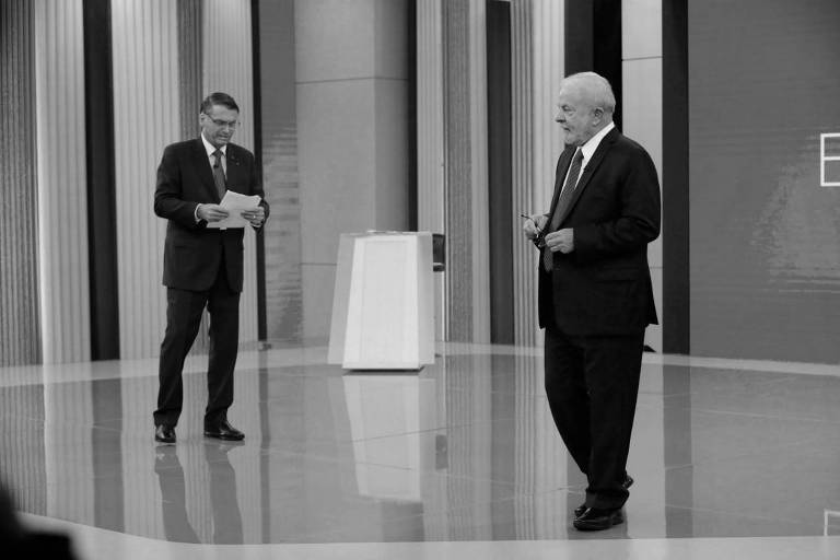 Bolsoaro, à esquerda, lê um texto em um papel, enquanto Lula, à direita da imagem, caminha olhando para uma câmera. Ambos vestem ternos pretos 