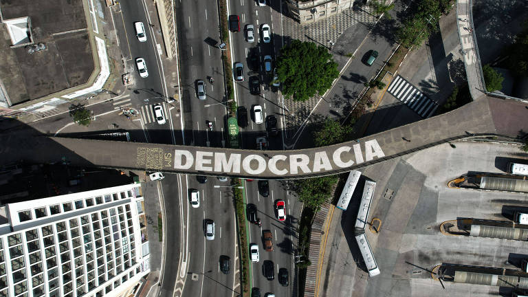 Imagem aérea mostra intervenção no asfalto no entorno do Terminal Bandeira, região central de São Paulo; em passarela escrito Democracia e movimento de ônibus e carros abaixo 