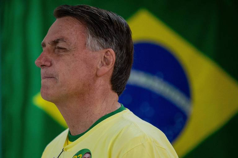 Bolsonaro, um homem branco, cabelos castanhos curtos e lisos, vestido com uma camiseta amarela com gola verde. Ele está de perfil, com os olhos e lábios cerrados, diante de uma bandeira do Brasil